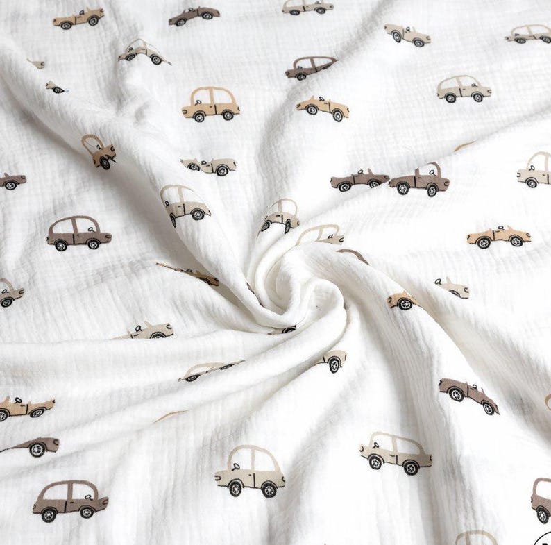 May include: White muslin fabric with a repeating pattern of small, tan and brown cars. The cars are in various styles, including sedans and trucks. The fabric is soft and has a crinkled texture, with a slight swirl in the center.