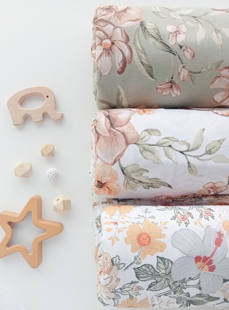 Puede incluir: Tres muselinas de tela floral plegadas en verde salvia, blanco y crema, con un elefante, una estrella y cuentas geom&eacute;tricas de madera. Las telas presentan grandes y detallados estampados florales en tonos melocot&oacute;n, rosa y gris.