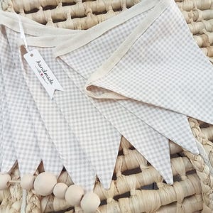 May include: A handmade beige and white gingham fabric bunting with wooden beads. The pennant flags are attached to a cream-coloured ribbon. A tag that says "handmade" is attached to the bunting.