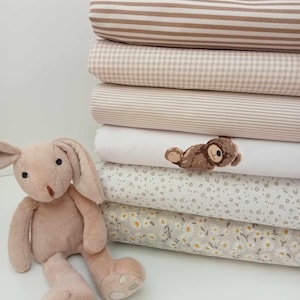 May include: A stack of six fabric swatches in various patterns and colours, including beige, white, and brown. The patterns include stripes, gingham, and floral prints. A small plush brown bear is embroidered on one of the swatches. A plush pink bunny sits in the foreground.