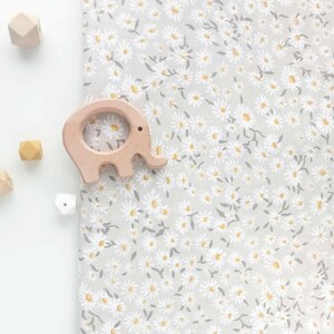Puede incluir: Un mordedor de beb&eacute; de madera con forma de elefante y varias cuentas de madera se colocan sobre una tela gris claro con un estampado floral de margaritas blancas. Las margaritas tienen centros amarillos y tallos grises.