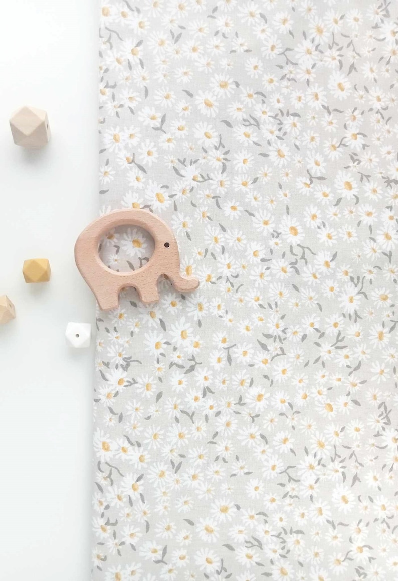 Puede incluir: Un mordedor de beb&eacute; de madera con forma de elefante y varias cuentas de madera se colocan sobre una tela gris claro con un estampado floral de margaritas blancas. Las margaritas tienen centros amarillos y tallos grises.