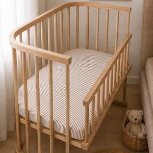 Peut inclure: Un berceau en bois clair avec des coins arrondis et des barreaux verticaux, doté d'un matelas recouvert d'un drap en tissu à rayures blanches et brunes. Le berceau présente une conception à côté ouvert en bois naturel lisse.