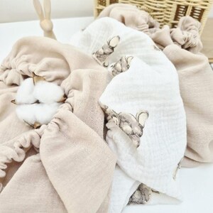 May include: A collection of soft, beige and white baby blankets and accessories. One white blanket features a repeating elephant pattern. A small wooden bunny and a woven basket are in the background. Cotton bolls are in the foreground.