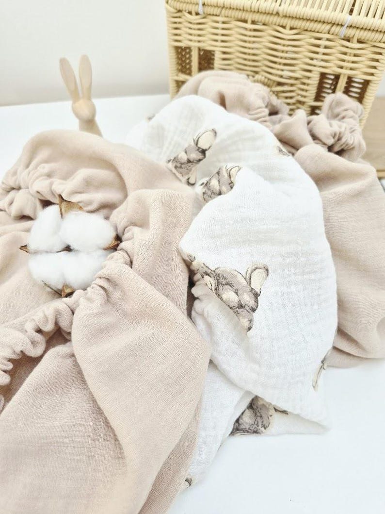 May include: A collection of soft, beige and white baby blankets and accessories. One white blanket features a repeating elephant pattern. A small wooden bunny and a woven basket are in the background. Cotton bolls are in the foreground.