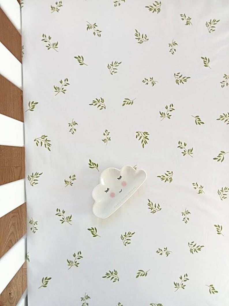 Puede incluir: S&aacute;bana blanca para cuna con un patr&oacute;n repetido de hojas verdes. Un plato blanco en forma de nube con una cara sonriente descansa sobre la s&aacute;bana. La s&aacute;bana est&aacute; junto a una cuna de madera.