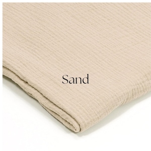 May include: A folded piece of sand-colored fabric with a textured, crinkled appearance. The word "Sand" is printed in black on the fabric. The fabric appears to be soft and lightweight, possibly a type of gauze or muslin.
