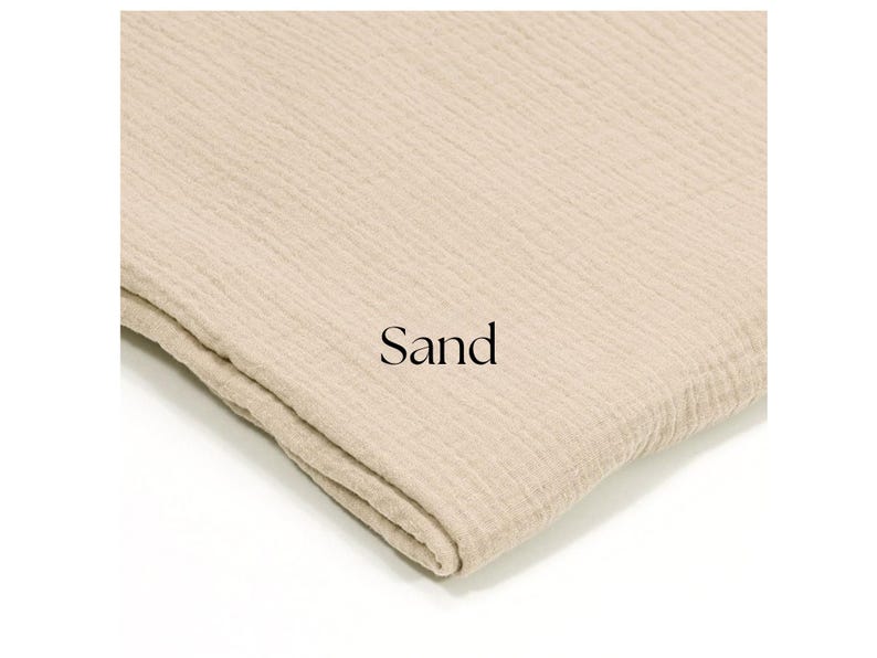 May include: A folded piece of sand-colored fabric with a textured, crinkled appearance. The word "Sand" is printed in black on the fabric. The fabric appears to be soft and lightweight, possibly a type of gauze or muslin.