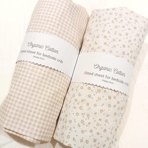 May include: Two rolled organic cotton fitted sheets for a bedside crib, each wrapped in a white paper band labeled Organic Cotton Fitted sheet for bedside crib Happy Dots. One sheet features a beige gingham pattern, the other a beige floral print.