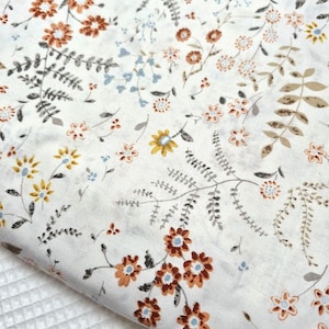 Puede incluir: Primer plano de una tela blanca con un estampado floral. El dise&ntilde;o presenta varias flores en tonos marr&oacute;n, naranja, amarillo y azul, junto con hojas y tallos grises y marrones. La tela est&aacute; doblada, revelando una superficie blanca texturizada.