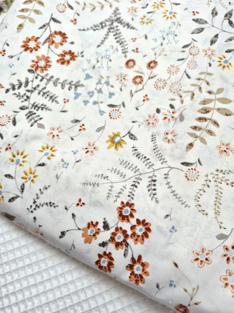 Puede incluir: Primer plano de una tela blanca con un estampado floral. El dise&ntilde;o presenta varias flores en tonos marr&oacute;n, naranja, amarillo y azul, junto con hojas y tallos grises y marrones. La tela est&aacute; doblada, revelando una superficie blanca texturizada.