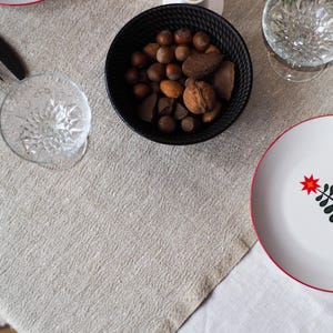 May include: A festive table setting with a white plate featuring a Christmas tree design with a red star. A black bowl holds nuts, and a clear glass and black fork are also visible. The tablecloth is a neutral color.