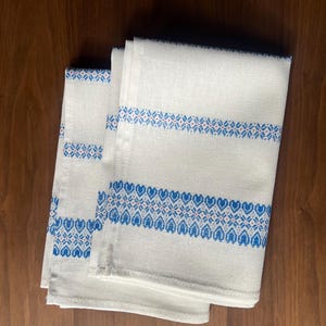 May include: Two folded off-white kitchen towels featuring horizontal bands of blue and red geometric patterns. The design includes repeating heart-shaped motifs and intricate cross-stitch style embroidery across the width of the fabric.