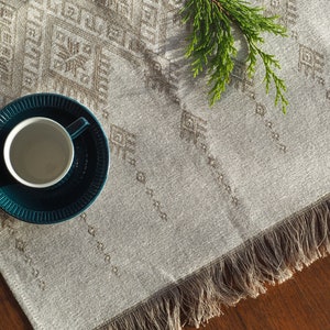May include: A teal teacup and saucer set on a patterned beige table runner with fringe. The runner has a geometric design and a sprig of greenery. The teacup is white inside.