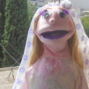 May include: A pink puppet dressed as a bride, wearing a veil with purple floral accents. The puppet has blonde hair, large eyes, and a purple mouth. The puppet is wearing a pink dress with a floral pattern.