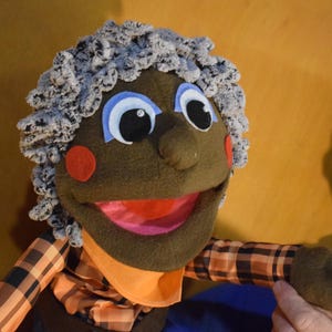 Op de afbeelding: Een handgemaakte pop met een bruin gezicht, grote blauwe ogen en grijs krullend haar. De pop heeft rode wangen en een wijd open mond. Ze draagt een geruit overhemd met een oranje kraag. De pop is ontworpen om door een hand te worden vastgehouden.
