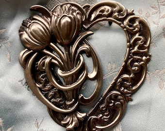 May include: A silver-toned metal heart-shaped ornament with a floral design. The ornament features two stylized tulips with leaves and stems, all intricately detailed.