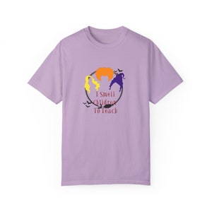 Peut inclure: Un t-shirt lavande avec un graphique de trois sorcières dans un cercle avec le texte "I Smell Children To Teach".