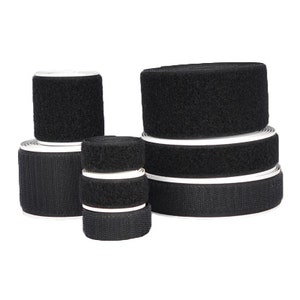 May include: Several rolls of black hook and loop tape, also known as Velcro, are stacked and arranged. The rolls vary in size, with the hook side facing up. The tape has a white backing.