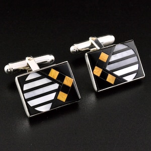 May include: A pair of square cufflinks with a silver-tone metal base. Each cufflink features a black background with a geometric design of white, gray, and gold-colored accents. The cufflinks have a classic, formal style.