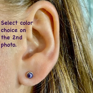 May include: A close-up of a person's ear with a small, round, purple and white gemstone earring. The earring is set in a silver setting.