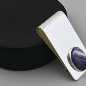 May include: A polished silver money clip with a rounded, oval-shaped, purple and white stone inlay. The clip has a simple, elegant design, perfect for holding cash and cards. The stone has a striped pattern.