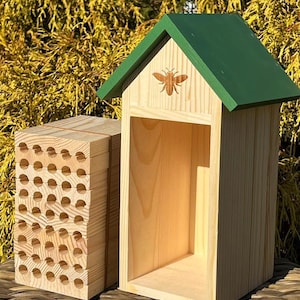 May include: A wooden bee house and a separate bee hotel. The bee house has a green roof and a bee-shaped cutout. The bee hotel is made of stacked wooden blocks with numerous holes. Both are made of light-colored wood.