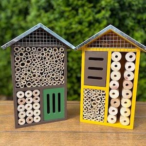 May include: Two wooden insect houses with metal roofs. One is brown with a green door and filled with bamboo tubes and drilled holes. The other is yellow with a brown section and filled with bamboo tubes and drilled holes.