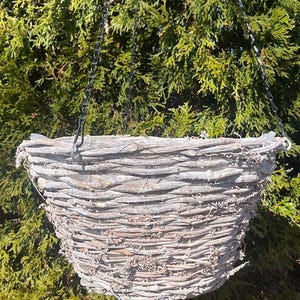 May include: A hanging basket made of woven branches, suspended by a metal chain. The basket has a natural, weathered appearance with a light gray color. The background is a dense, green hedge.