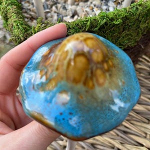 K&ouml;nnte beinhalten: Ein handgefertigter Keramikpilz mit blauer und brauner Glasur. Der Pilz wird in einer Hand gehalten, mit einem wei&szlig;en Stiel und einem moosbedeckten Sockel im Hintergrund. Der Pilz ist ein dekorativer Artikel.