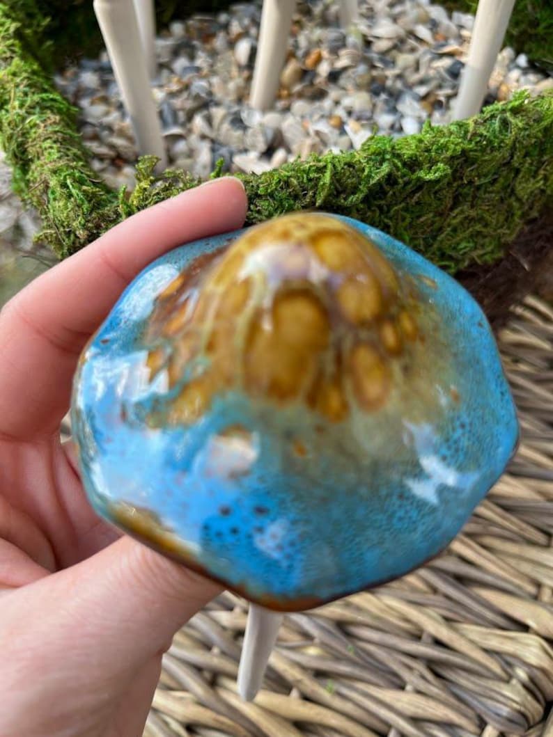K&ouml;nnte beinhalten: Ein handgefertigter Keramikpilz mit blauer und brauner Glasur. Der Pilz wird in einer Hand gehalten, mit einem wei&szlig;en Stiel und einem moosbedeckten Sockel im Hintergrund. Der Pilz ist ein dekorativer Artikel.