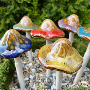 K&ouml;nnte beinhalten: Eine Sammlung von Keramik-Pilzskulpturen mit gl&auml;nzenden, farbenfrohen Kappen in Blau-, Rot-, Gold- und T&uuml;rkist&ouml;nen. Jeder Pilz hat einen wei&szlig;en Stiel und ist in einem Bett aus Moos und kleinen Steinen platziert.