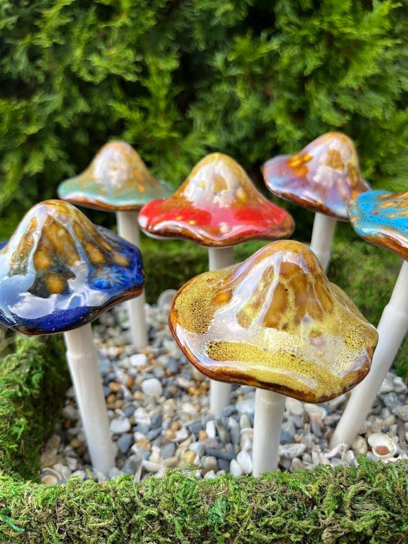K&ouml;nnte beinhalten: Eine Sammlung von Keramik-Pilzskulpturen mit gl&auml;nzenden, farbenfrohen Kappen in Blau-, Rot-, Gold- und T&uuml;rkist&ouml;nen. Jeder Pilz hat einen wei&szlig;en Stiel und ist in einem Bett aus Moos und kleinen Steinen platziert.