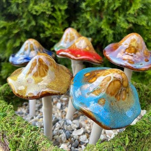 K&ouml;nnte beinhalten: Eine Ansammlung von Keramik-Pilzskulpturen mit glasierten Kappen in verschiedenen Farben, darunter Blau, Rot und Gold. Die Pilze sind in einem Bett aus Moos und kleinen Steinen angeordnet, vor einem Hintergrund aus gr&uuml;nem Laub.