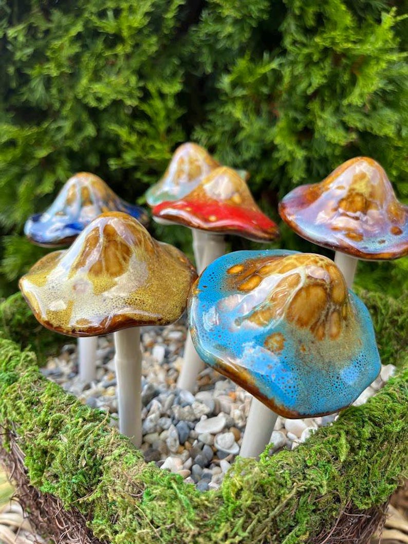 K&ouml;nnte beinhalten: Eine Ansammlung von Keramik-Pilzskulpturen mit glasierten Kappen in verschiedenen Farben, darunter Blau, Rot und Gold. Die Pilze sind in einem Bett aus Moos und kleinen Steinen angeordnet, vor einem Hintergrund aus gr&uuml;nem Laub.