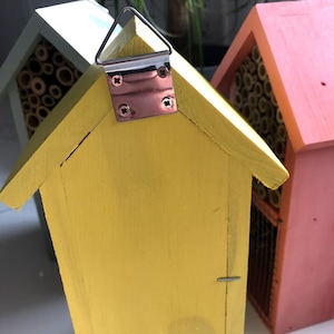 May include: A yellow wooden bee house with a metal hanging loop. The bee house has a small opening on the front and a textured surface. The bee house is sitting on a white surface.