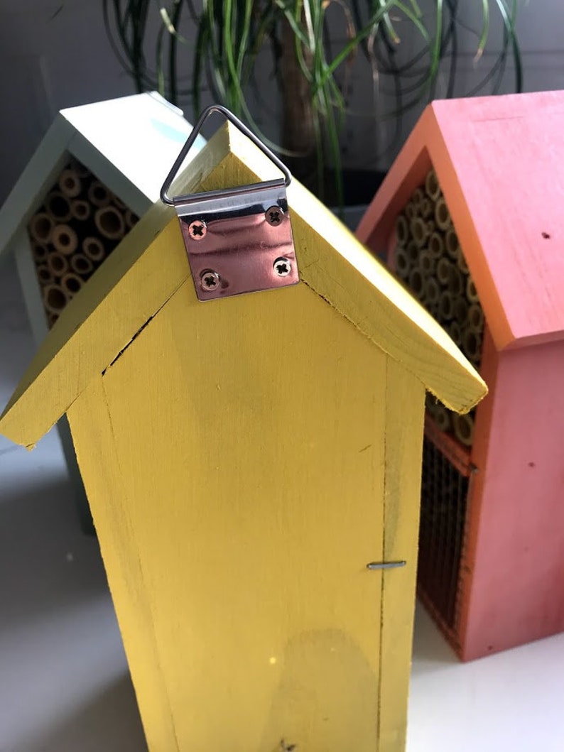 May include: A yellow wooden bee house with a metal hanging loop. The bee house has a small opening on the front and a textured surface. The bee house is sitting on a white surface.
