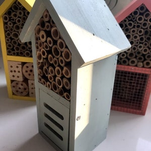 May include: Three insect houses made of wood with different colored paint. The house on the left is yellow, the middle house is light blue, and the house on the right is red. Each house has a variety of compartments for insects to live in.