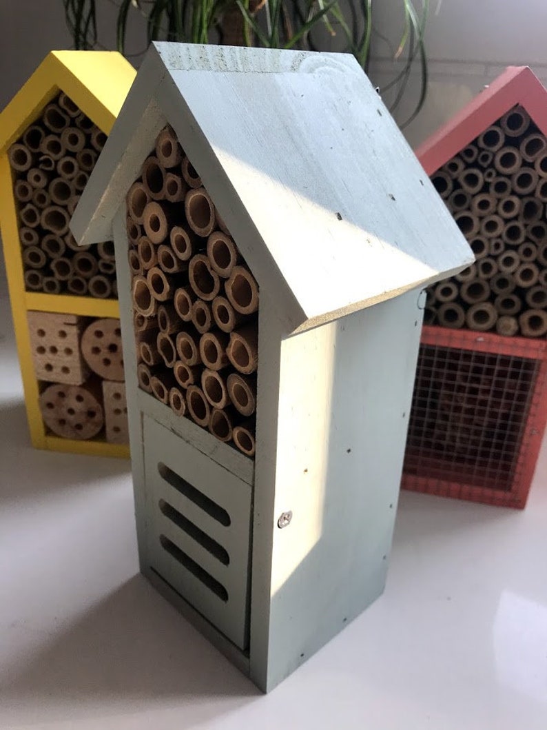 May include: Three insect houses made of wood with different colored paint. The house on the left is yellow, the middle house is light blue, and the house on the right is red. Each house has a variety of compartments for insects to live in.