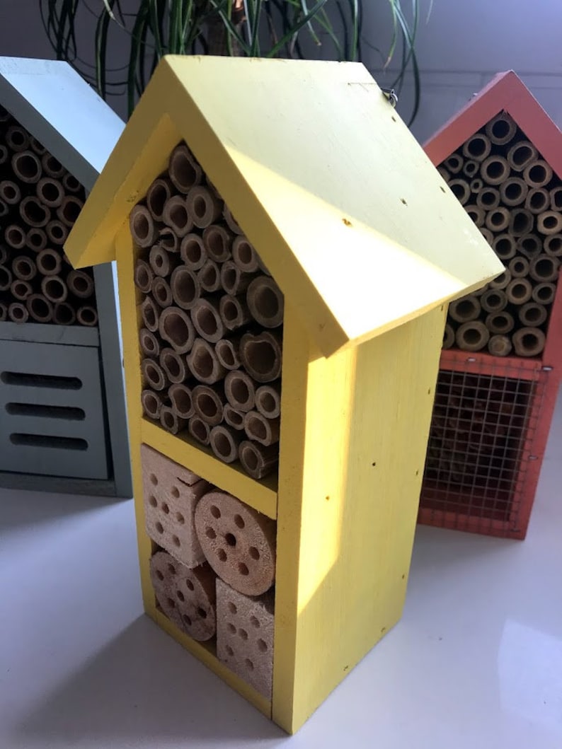 May include: A yellow wooden insect house with multiple compartments for different types of insects. The house has a variety of materials, including bamboo, wood, and pine cones.