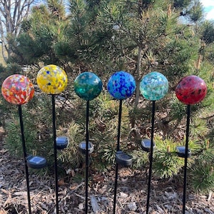 May include: Six colorful glass globe solar garden lights. Each globe is a different color, including red, yellow, blue, and green, with a speckled design. The lights are mounted on black metal stakes and are set in a garden bed.