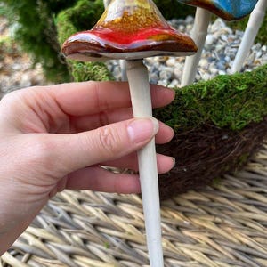 K&ouml;nnte beinhalten: Ein keramischer Gartenstecker in Pilzform mit einer rot, braun und gelb glasierten Kappe. Der Stecker wird in einer Hand gehalten, mit anderen bunten Pilzsteckern im Hintergrund. Die Pilzkappen haben eine gl&auml;nzende Oberfl&auml;che und sind auf einem hellen Holzstab befestigt.