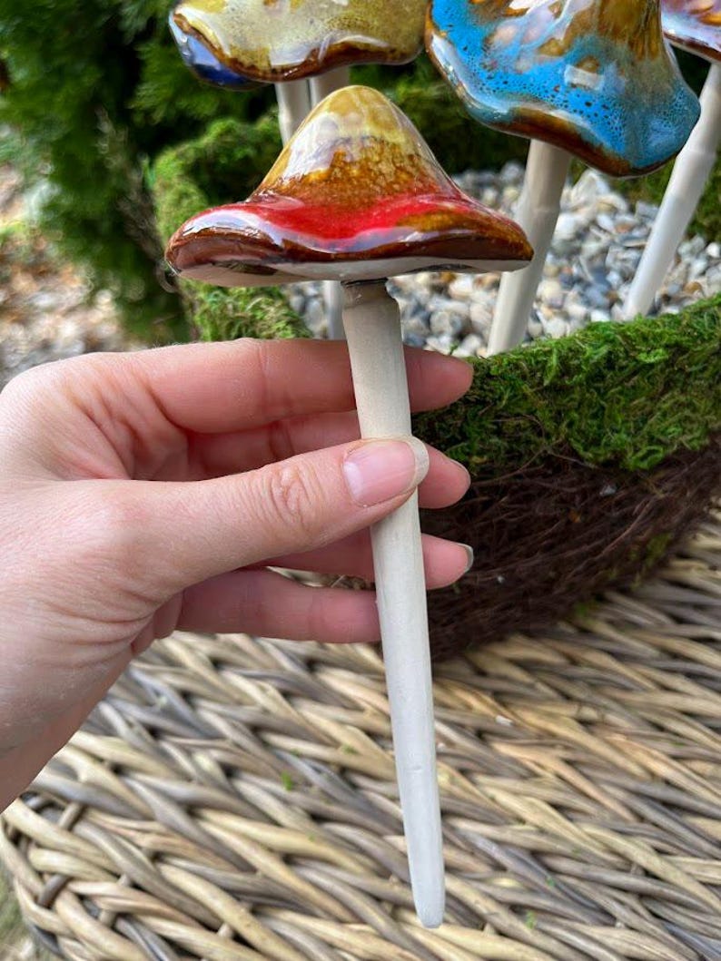 K&ouml;nnte beinhalten: Ein keramischer Gartenstecker in Pilzform mit einer rot, braun und gelb glasierten Kappe. Der Stecker wird in einer Hand gehalten, mit anderen bunten Pilzsteckern im Hintergrund. Die Pilzkappen haben eine gl&auml;nzende Oberfl&auml;che und sind auf einem hellen Holzstab befestigt.