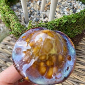 K&ouml;nnte beinhalten: Nahaufnahme eines Keramikpilzes mit gl&auml;nzend glasierter Oberfl&auml;che. Die Pilzkappe weist Braunt&ouml;ne, Gold und Blau auf und hat eine strukturierte Optik. Der Pilz ist Teil eines dekorativen Gartenornaments.