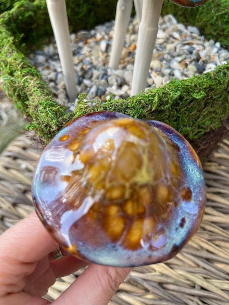 K&ouml;nnte beinhalten: Nahaufnahme eines Keramikpilzes mit gl&auml;nzend glasierter Oberfl&auml;che. Die Pilzkappe weist Braunt&ouml;ne, Gold und Blau auf und hat eine strukturierte Optik. Der Pilz ist Teil eines dekorativen Gartenornaments.