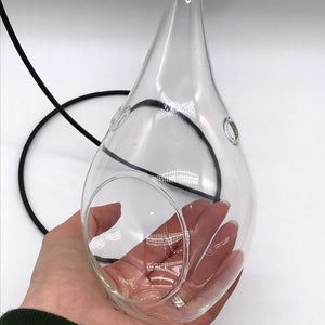Puede incluir: Terrario de vidrio transparente en forma de l&aacute;grima, suspendido por un cord&oacute;n negro y cuerda natural. El terrario tiene una gran abertura y dos peque&ntilde;os agujeros. El objeto se sostiene en una mano sobre un fondo blanco.
