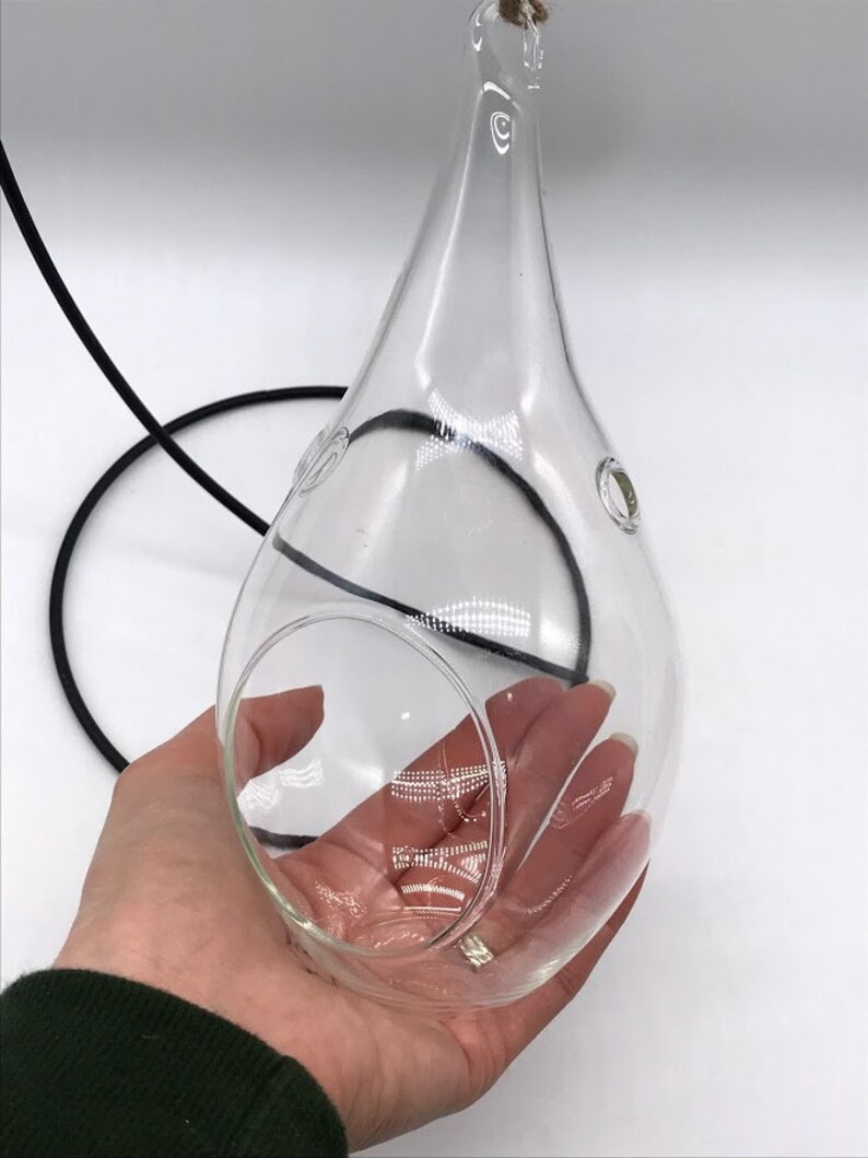 Puede incluir: Terrario de vidrio transparente en forma de l&aacute;grima, suspendido por un cord&oacute;n negro y cuerda natural. El terrario tiene una gran abertura y dos peque&ntilde;os agujeros. El objeto se sostiene en una mano sobre un fondo blanco.