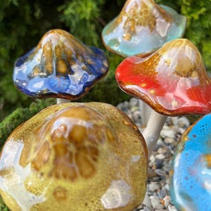 K&ouml;nnte beinhalten: Eine Sammlung von Keramik-Pilzskulpturen mit farbigen, glasierten Kappen. Die Pilze weisen eine Vielzahl von Farben auf, darunter Blau, Rot, Gelb und T&uuml;rkis, mit wei&szlig;en Stielen. Die Skulpturen sind vor einem Hintergrund aus Gr&uuml;n und kleinen Steinen platziert.