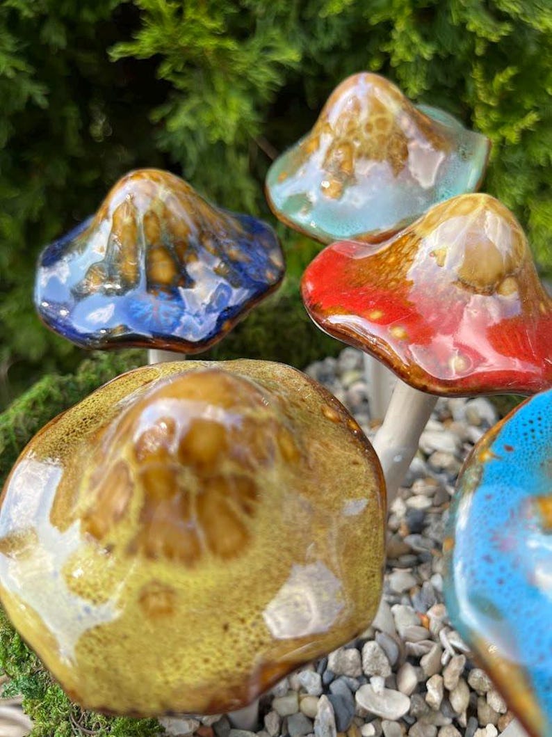K&ouml;nnte beinhalten: Eine Sammlung von Keramik-Pilzskulpturen mit farbigen, glasierten Kappen. Die Pilze weisen eine Vielzahl von Farben auf, darunter Blau, Rot, Gelb und T&uuml;rkis, mit wei&szlig;en Stielen. Die Skulpturen sind vor einem Hintergrund aus Gr&uuml;n und kleinen Steinen platziert.