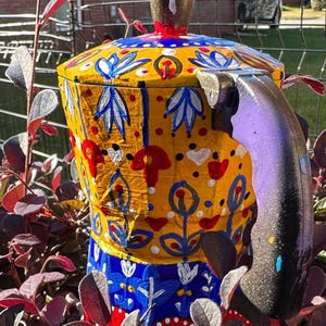 May include: A hand-painted, decorative teapot with a lid and handle. The teapot features a vibrant design with yellow, blue, red, and white floral patterns. The handle is purple and the lid has a gold-colored knob. The teapot is surrounded by red and green foliage.